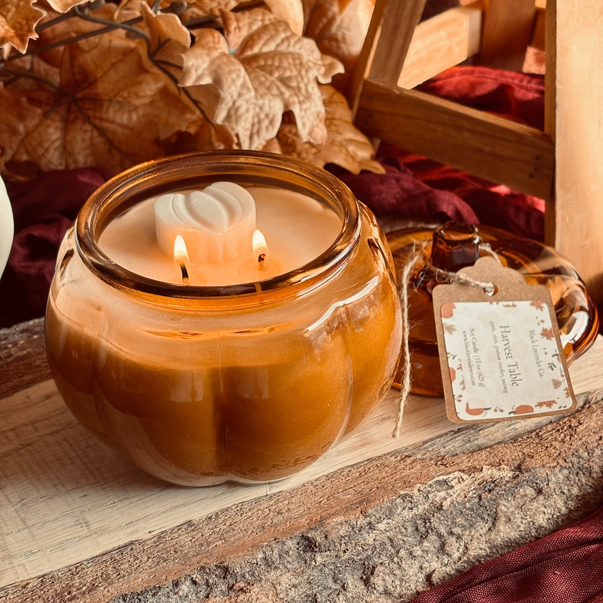 A pumpkin-shaped amber glass candle with two lit wicks sits on a rustic wooden surface, surrounded by dried autumn leaves, red fabric, and a small hanging tag.