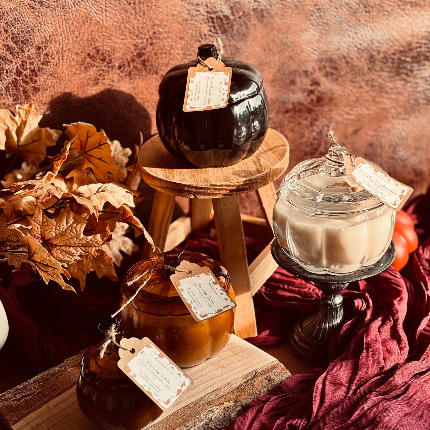 Three seasonal scented candles in decorative containers styled with autumn props. The set includes a black candle in a pumpkin-shaped vessel on a wooden stool, an amber glass jar with a brown candle, and a cream-colored candle in a clear glass apothecary jar, all with handwritten paper tags.