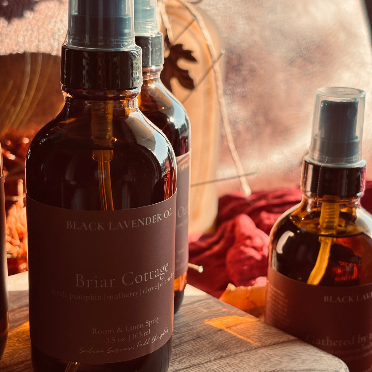 Three amber glass spray bottles with dark brown labels from Black Lavender Co., including Briar Cottage, arranged on a wooden surface with a warm, rustic background.