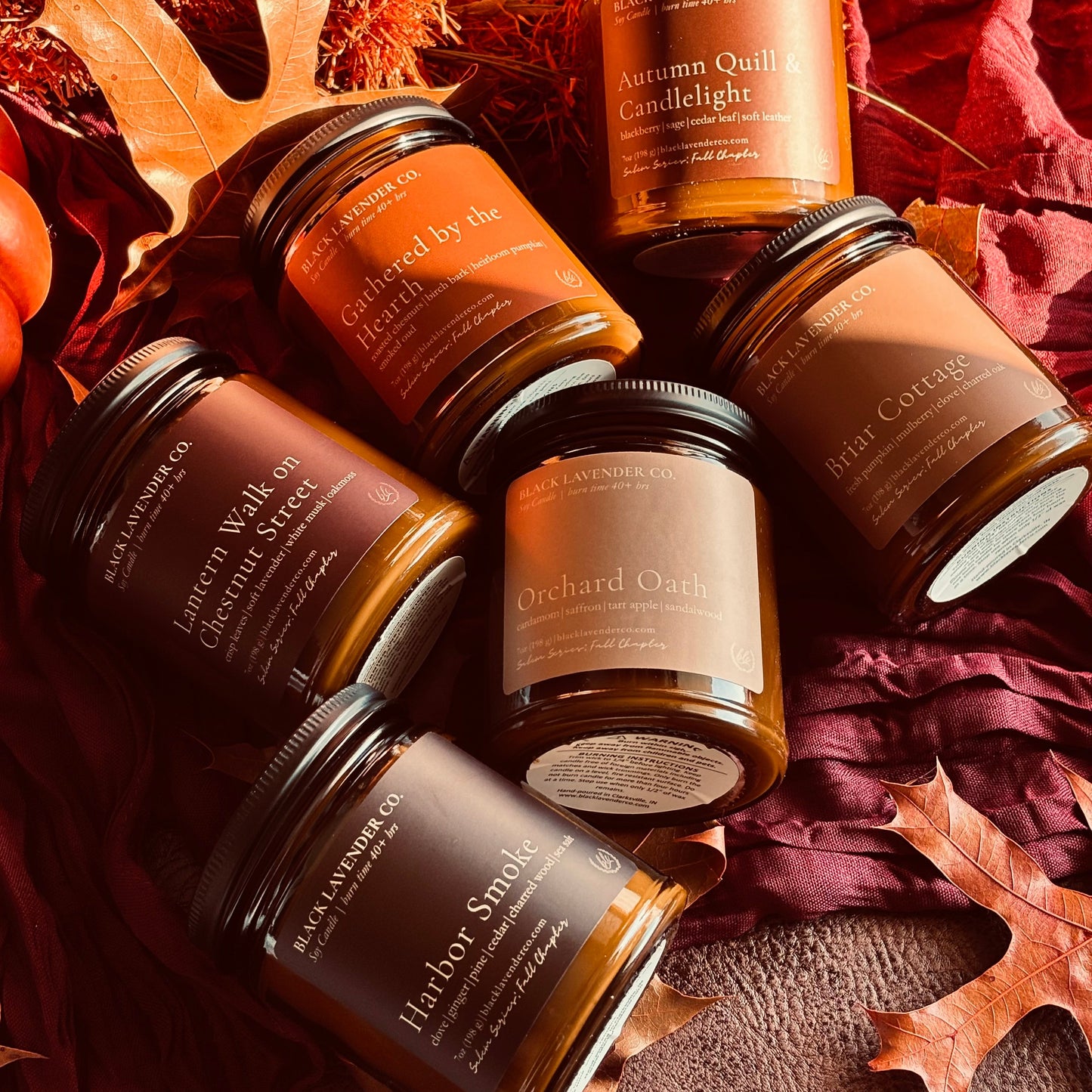 Six amber glass jar candles from Black Lavender Co. are arranged in a flat lay with autumn-themed props including dried orange leaves, red fabric, and a pumpkin.