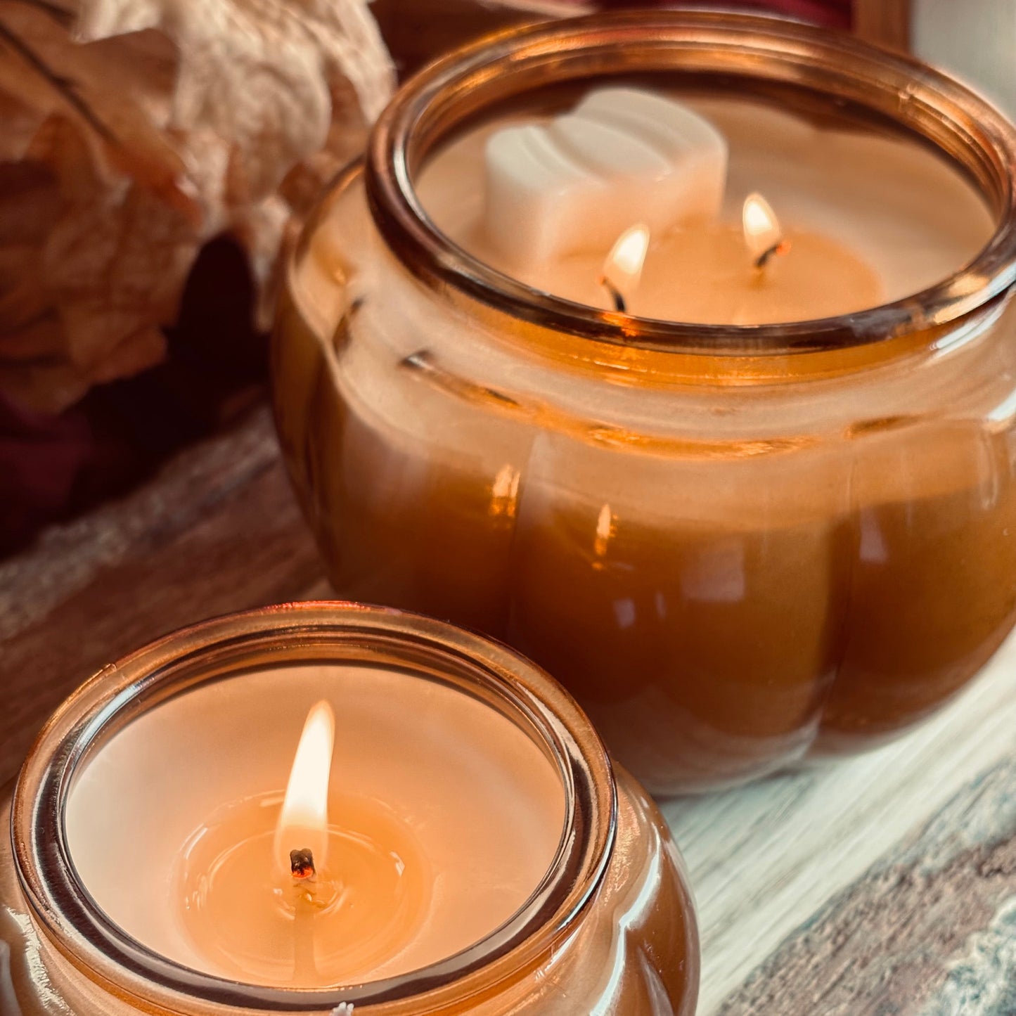 Two lit amber glass jar candles with white wax are arranged on a rustic wooden surface, with dried leaves in the background.