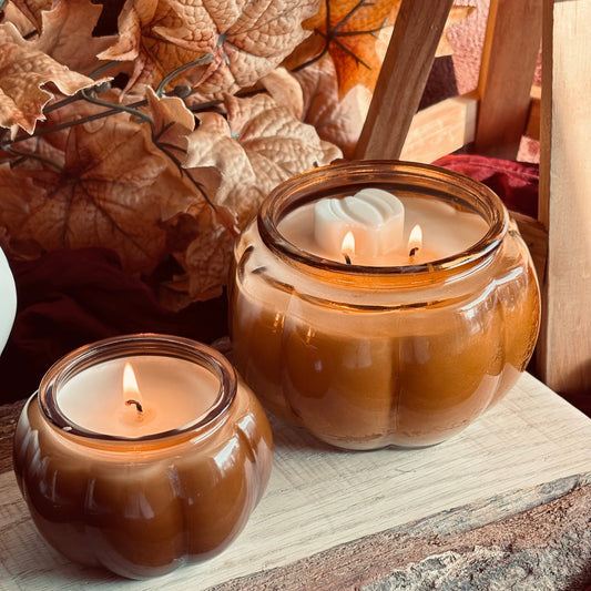 Two lit amber glass jar candles with a pumpkin-like shape, styled on a rustic stone surface with dried autumn leaves and burgundy fabric in the background.