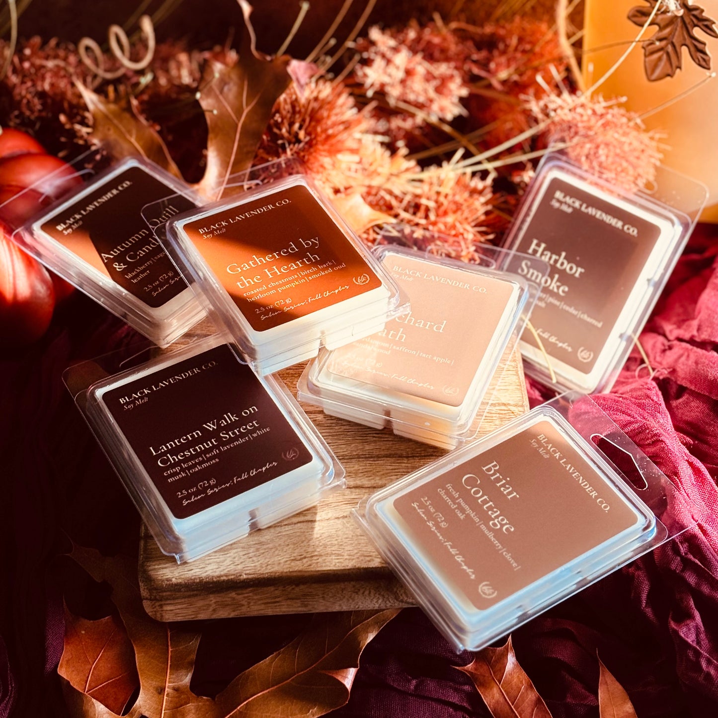 six Black Lavender Co. wax melts in clear clamshell packaging are arranged on a wooden board with autumn leaves and dried flowers in the background.