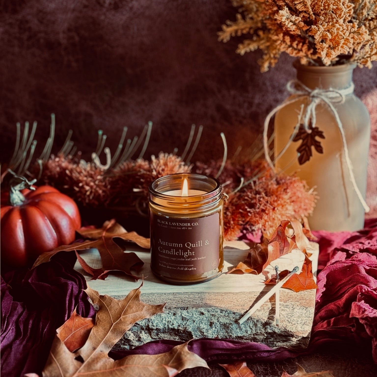 A lit candle in a amber glass jar sits on a rustic wood slab surrounded by autumn leaves, dried flowers, and a small red pumpkin. The candle label reads 'Autumn Quill & Candlelight' and shows the brand 'Black Lavender Co.