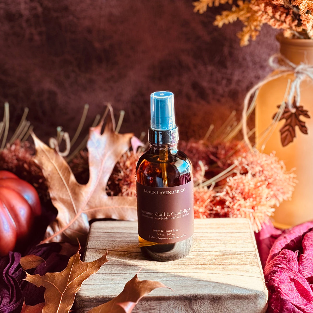 A bottle of Black Lavender Co room spray with a black spray top and amber glass, displayed on a wooden board surrounded by autumn leaves, a pumpkin, and dried flowers.
