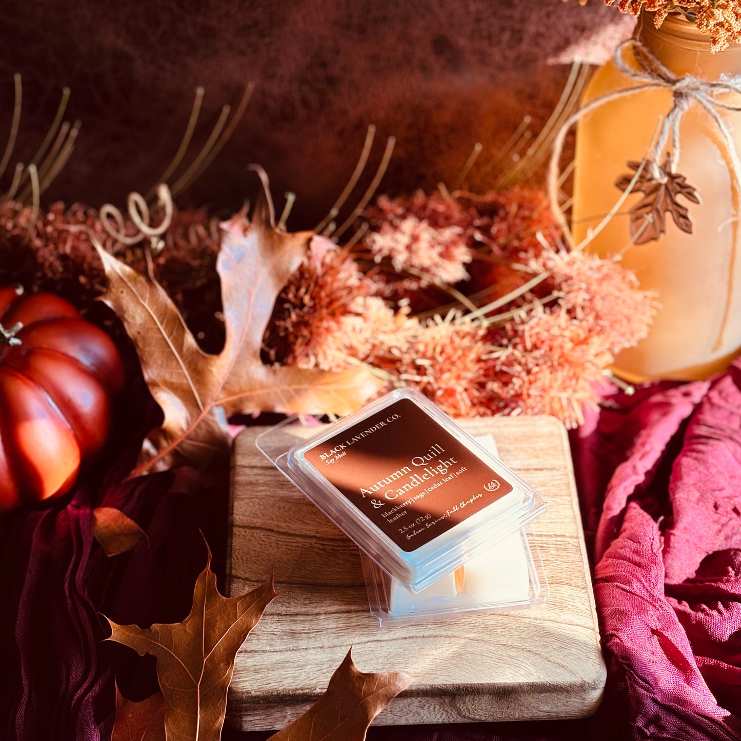 A wax melt package labeled 'Autumn Quill & Candlelight' by Black Lavender Co sits on a wooden block surrounded by autumn-themed props including dried leaves, a small pumpkin, and a vase