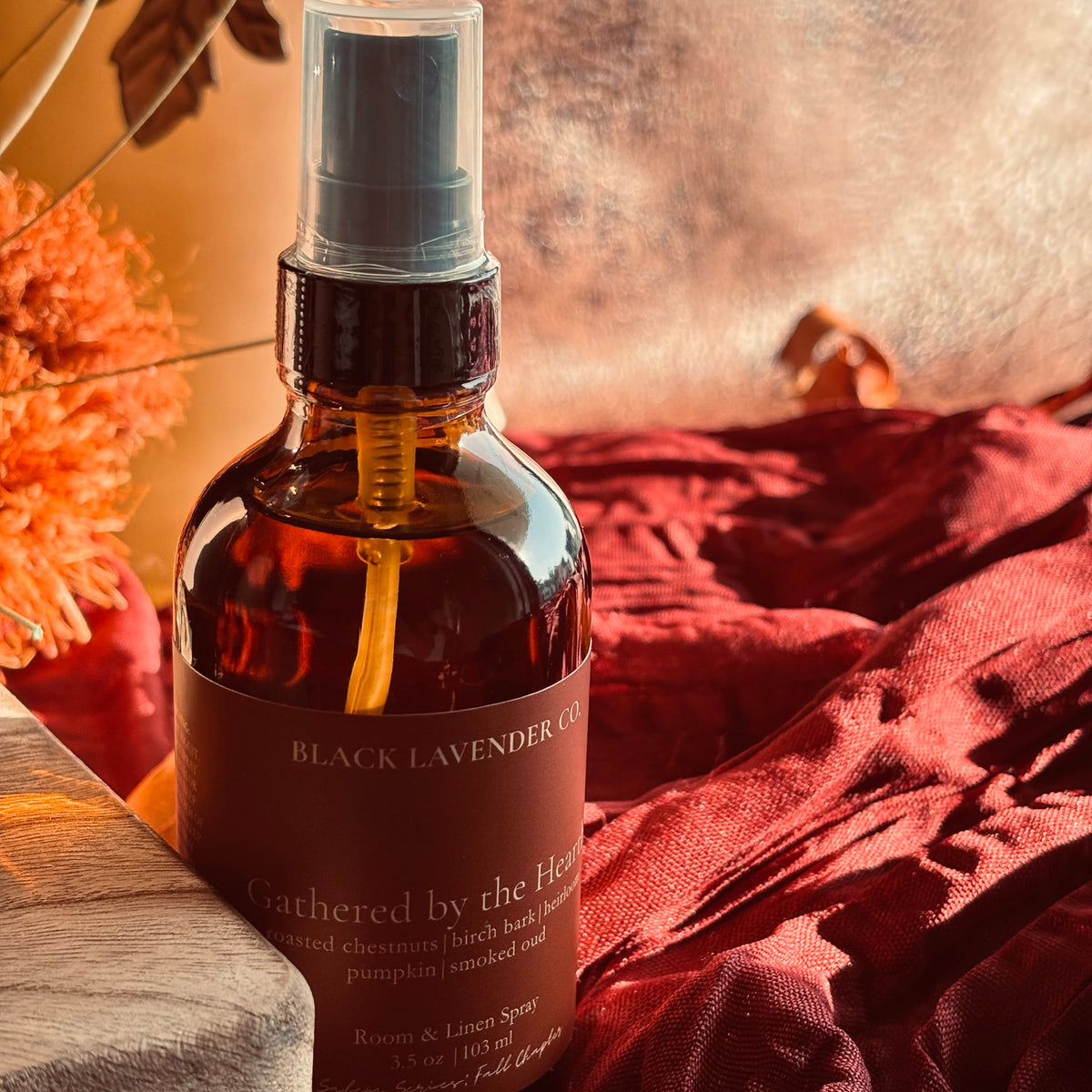 An amber glass spray bottle labeled 'Black Lavender Gathered by the Hearth' with a black spray top, styled on red fabric with dried orange flowers and a wooden block in warm autumn lighting.