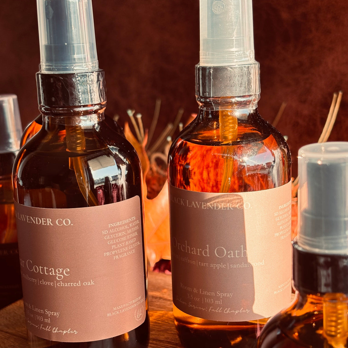 Three amber glass spray bottles with warm autumn color labels arranged on a wooden surface against a warm brown background.