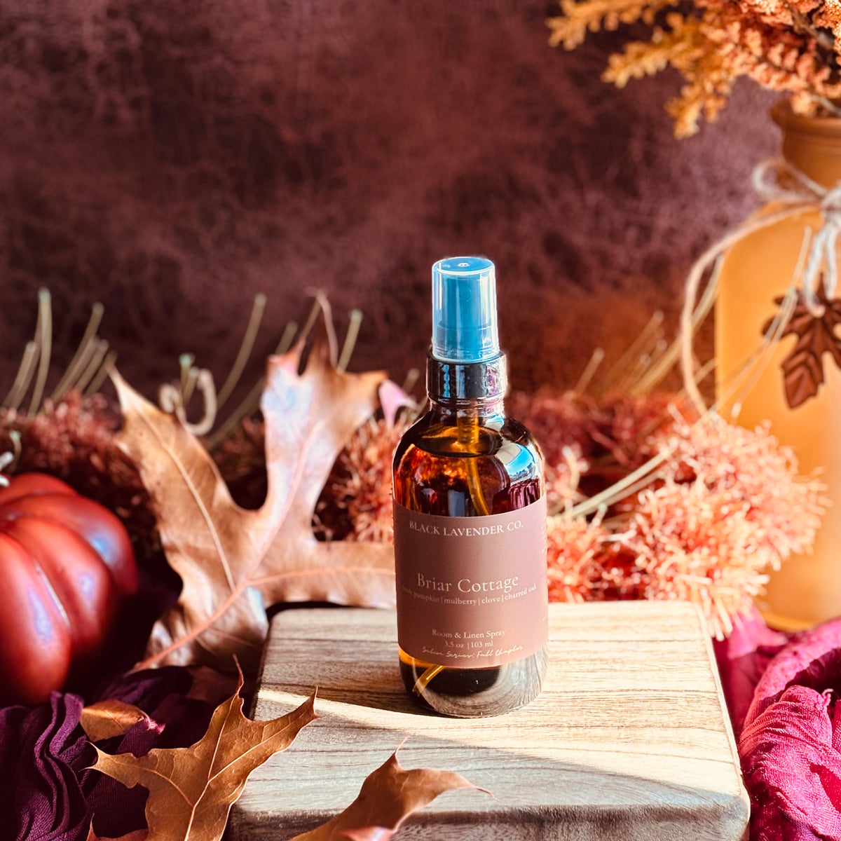 A bottle of Black Lavender Co. Briar COttage Room Spray displayed on a wooden block surrounded by autumn leaves, a small pumpkin, and dried floral decor.