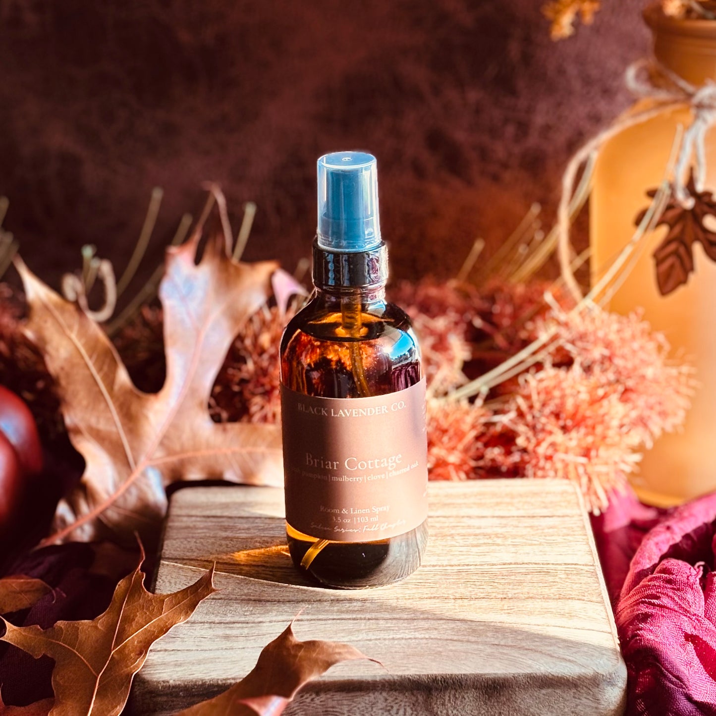 A bottle of Briar Cottage room and linen spray sits on a wooden block surrounded by autumn leaves, dried flowers, and a warm-toned decorative jar.
