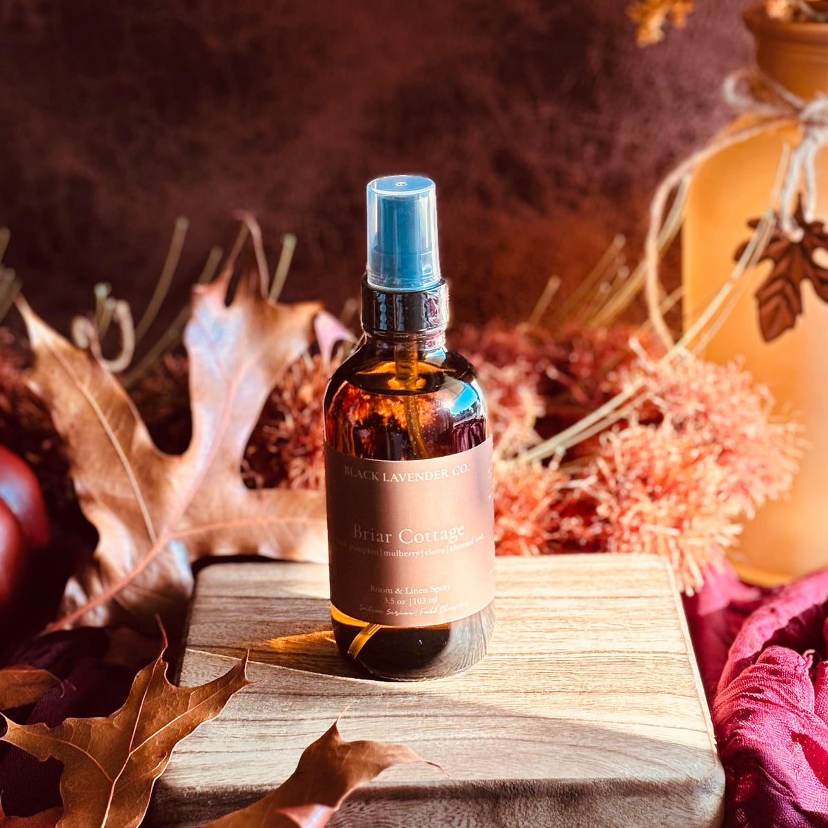 A bottle of Briar Cottage room and linen spray sits on a wooden block surrounded by autumn leaves, dried flowers, and a warm-toned decorative jar.