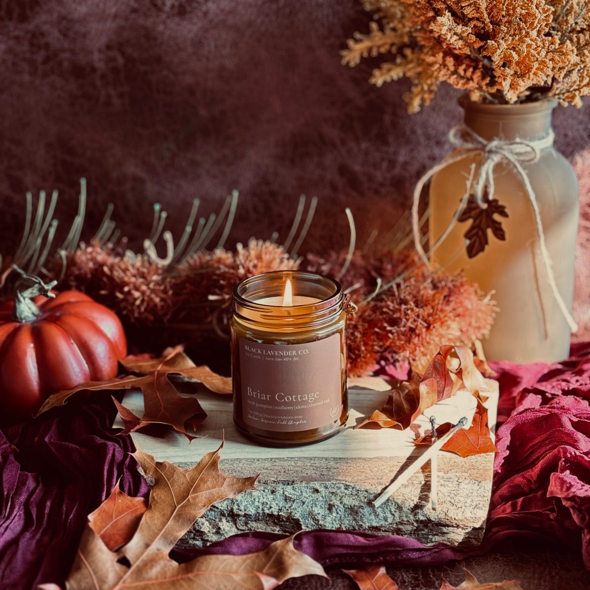 amber glass jar candle labeled Briar Cottage sits on a rustic wooden slab with autumn foliage