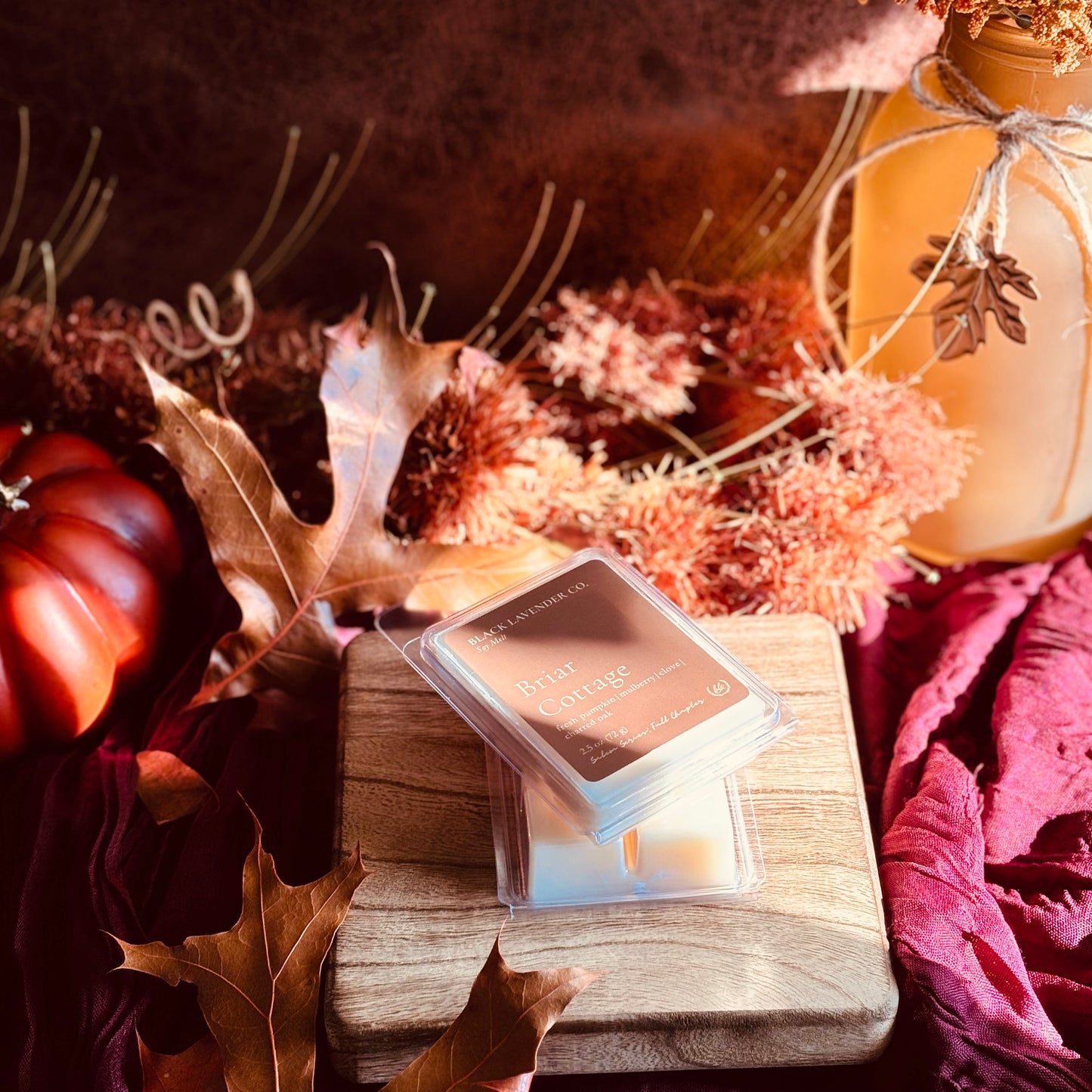 A wax melt package labeled 'Briar cottage' sits on a wooden block surrounded by autumn props including dried leaves, a small pumpkin, and a fabric surface in warm fall tones.