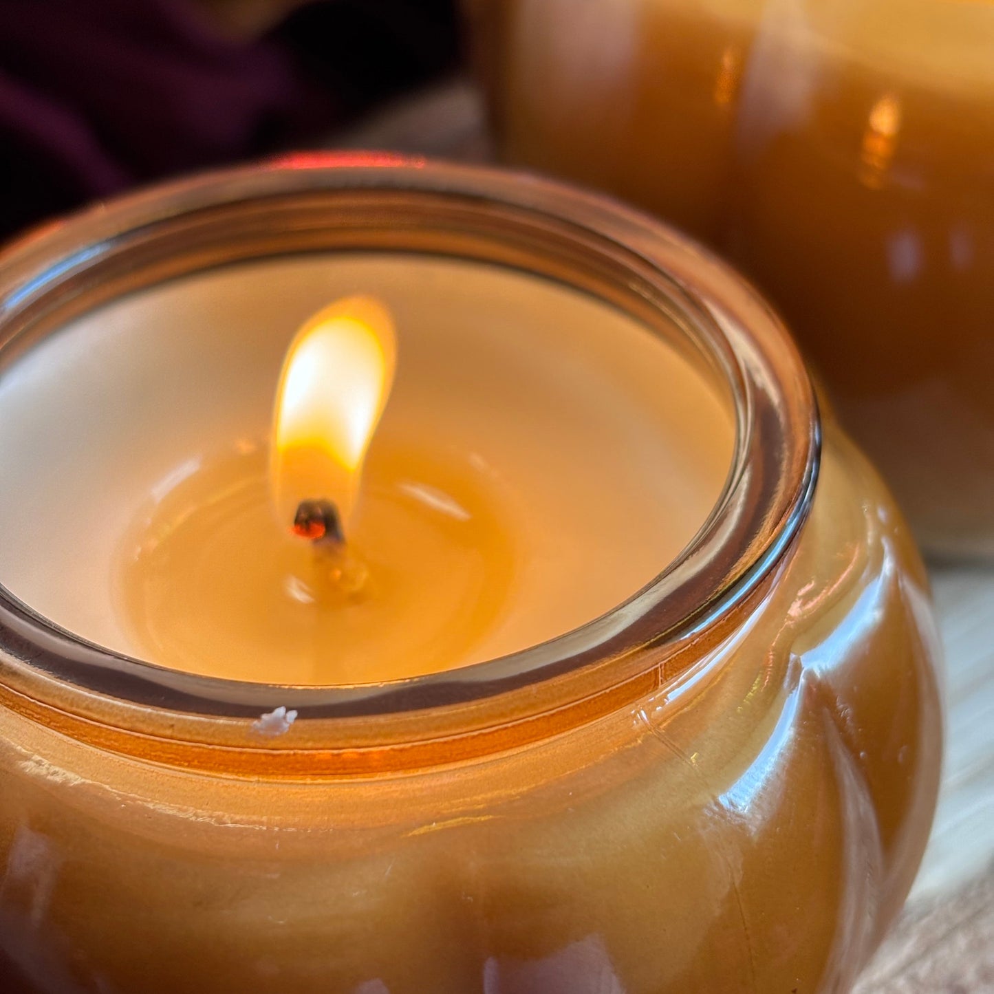 A close-up of a lit amber glass jar candle with a warm yellow flame, with another similar candle blurred in the background.