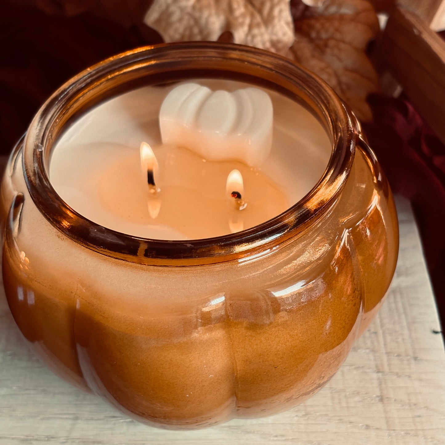 A lit two-wick candle in a translucent amber glass jar with a white pumpkin-shaped wax insert, set on a rustic wooden surface with autumn leaves in the background.