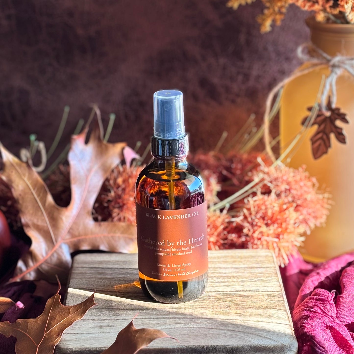 A bottle of Black Knavender Co 'Gathered by the Heart' room spray sits on a wooden block surrounded by autumn leaves and dried flowers.