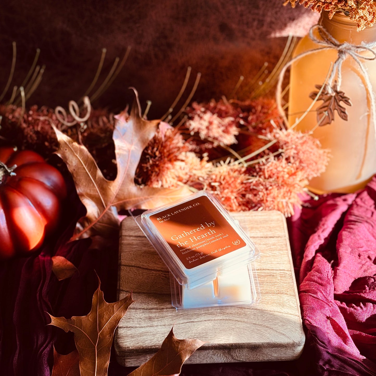 A small clear plastic jar candle labeled 'Gathered by the Hearth' sits on a wooden block in an autumn-themed styled scene with dried leaves, a small pumpkin, and a mason jar.