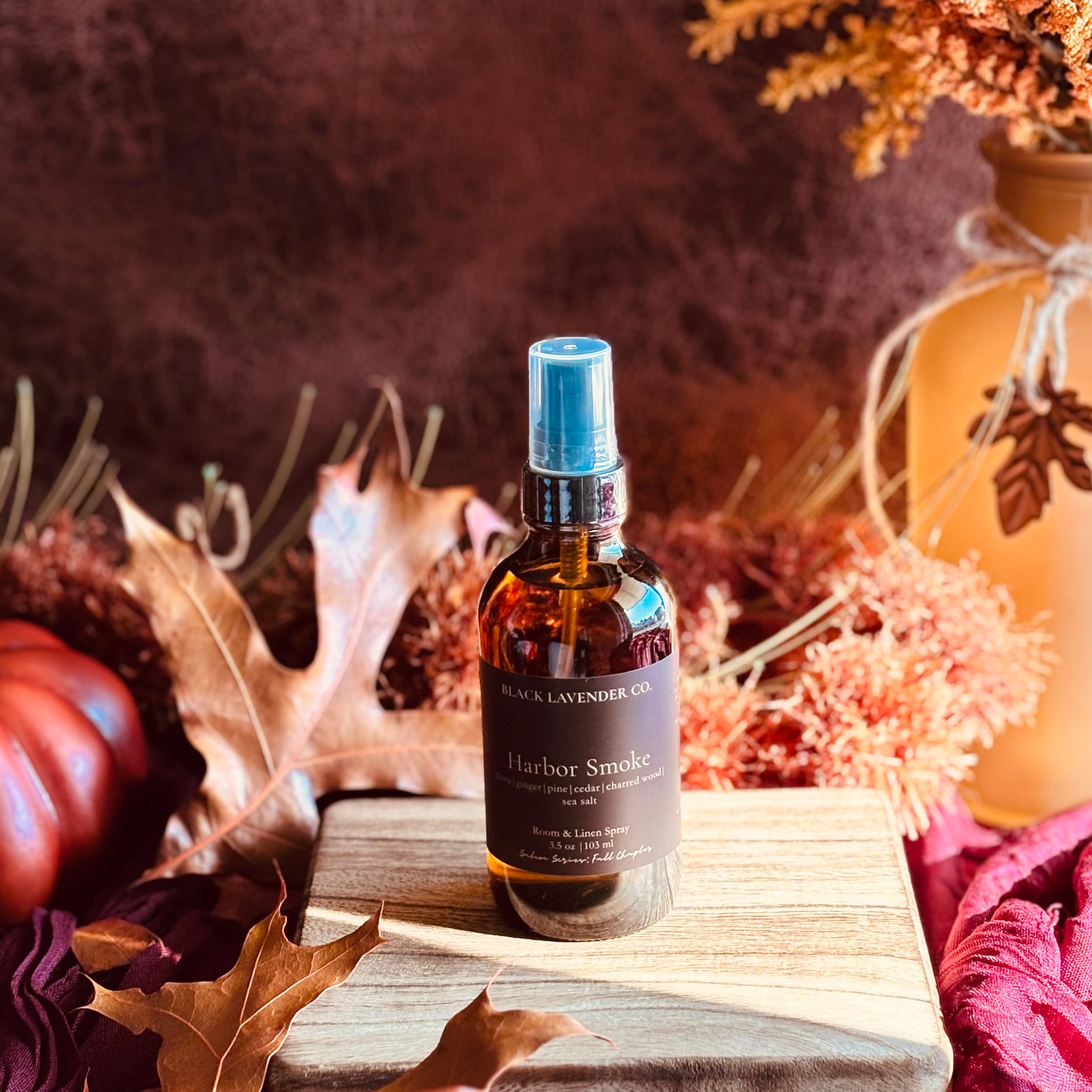 A bottle of Harbor Smoke room spray by Black Lavender Co. sits on a wooden block surrounded by autumn leaves, a pumpkin, and dried flowers in a warm, rustic setting.