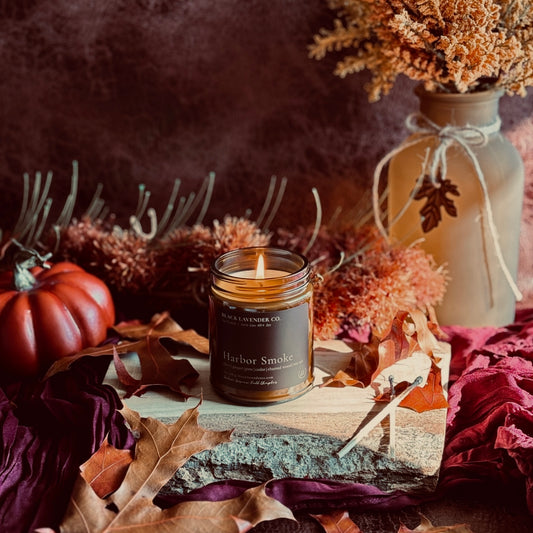A lit candle in a amber glass jar with a dark label reading 'Harbor Smoke' and 'Black Lavender Co.' sits on a rustic wood slab surrounded by autumn leaves, dried flowers, a small pumpkin, and a decorative vase against a dark textured background.