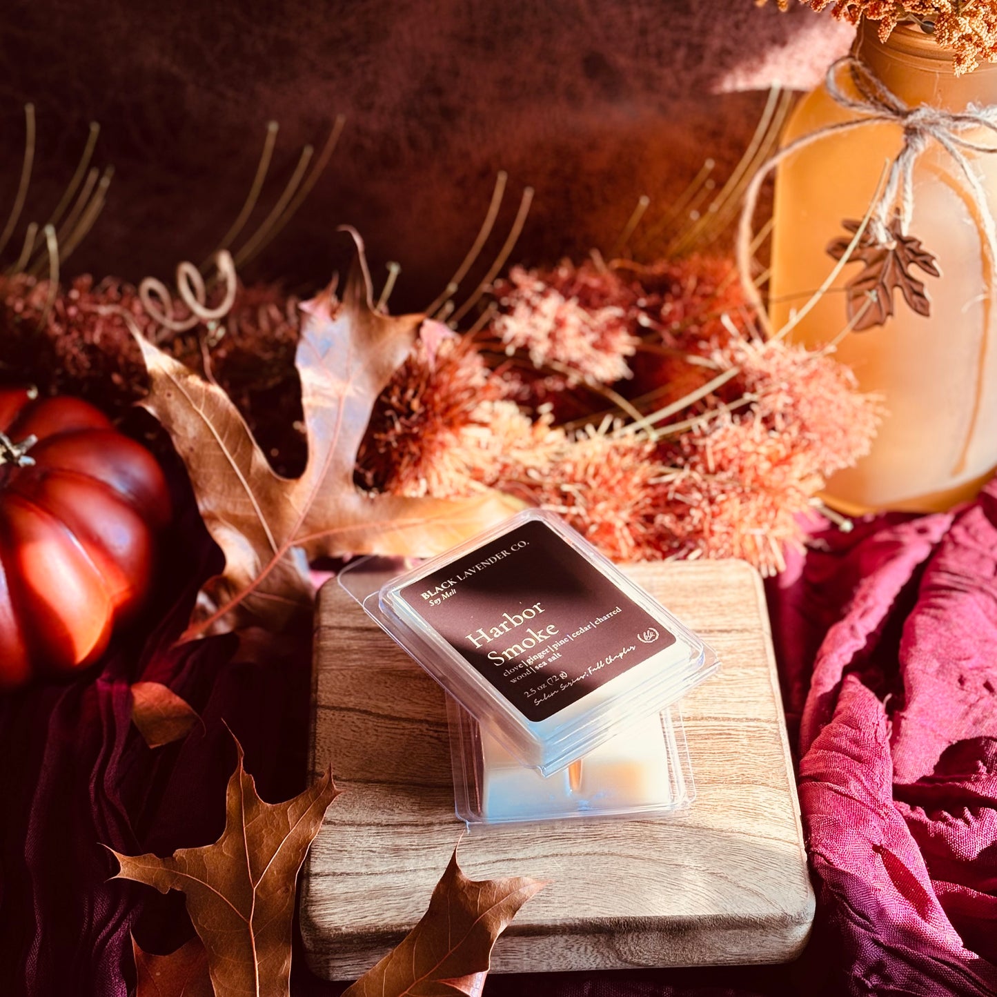 A wax melt package labeled 'Harbor Smoke' by Black Lavender Co. sits on a wooden block surrounded by autumn props including dried leaves, a small pumpkin, and a mason jar in warm fall colors.