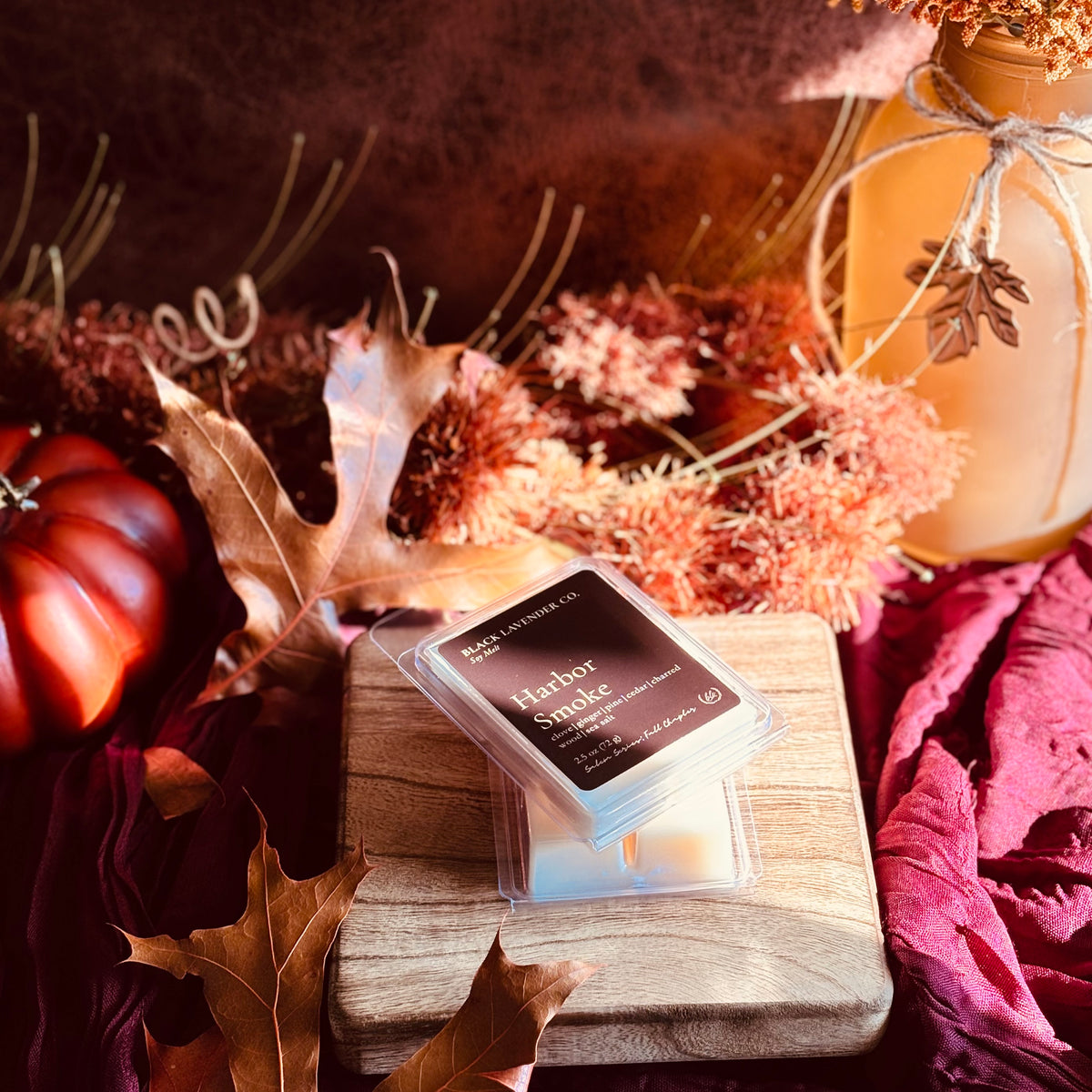 A wax melt package labeled 'Harbor Smoke' by Black Lavender Co. sits on a wooden block surrounded by autumn props including dried leaves, a small pumpkin, and a mason jar in warm fall colors.