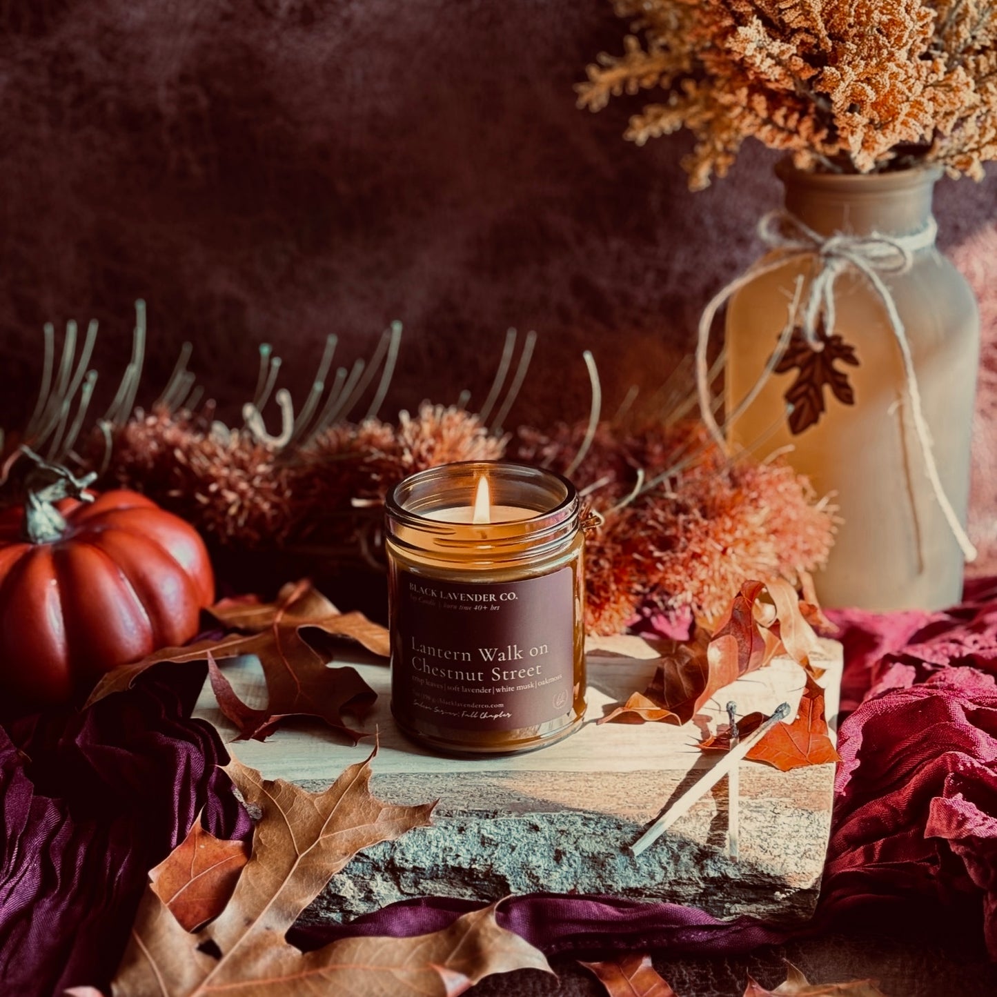 A lit candle in a amber glass jar sits on a rustic wood slab surrounded by autumn leaves, dried flowers, and a small pumpkin. A beige vase with twine and dried stems stands in the background against a dark brown backdrop.