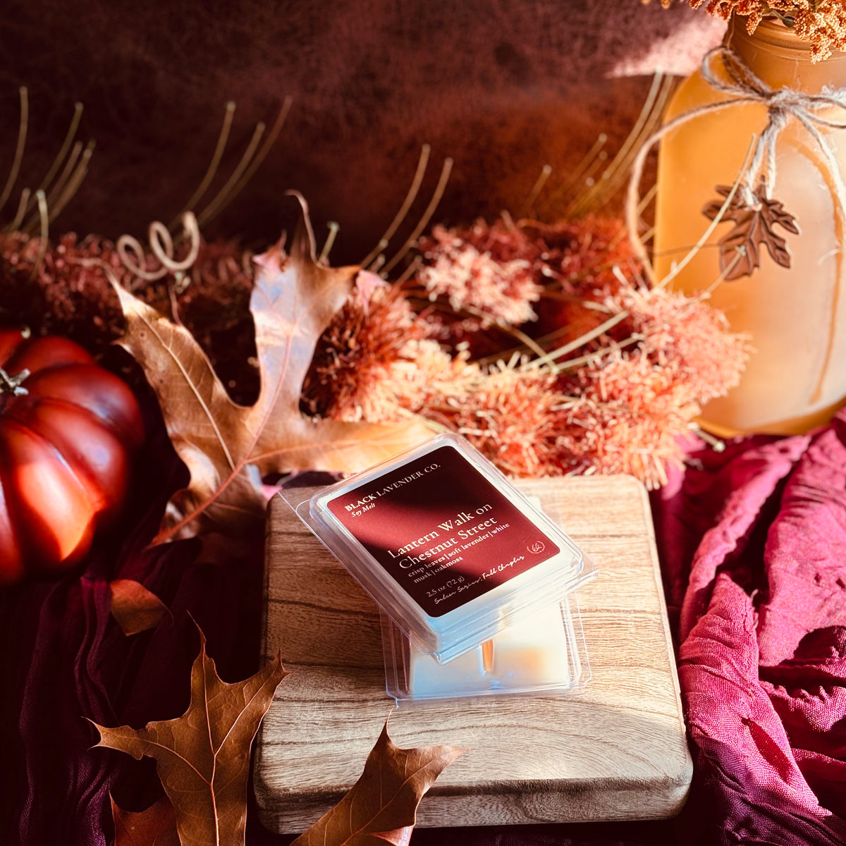 A packaged wax melt or scented melt product labeled 'Lantern Walk on Chestnut Street' sits on a wooden block surrounded by fall-themed props including dried leaves, a small pumpkin, and a mason jar in warm autumn colors.