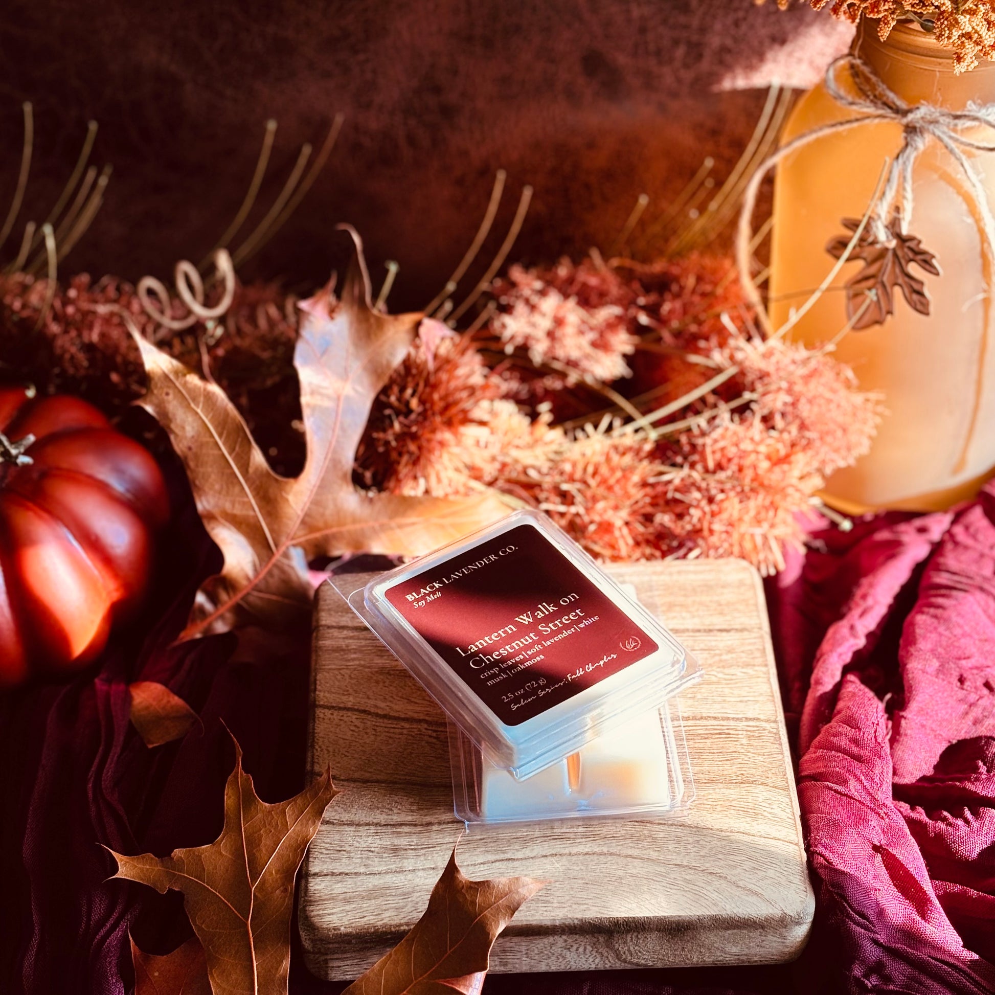 A packaged wax melt or scented melt product labeled 'Lantern Walk on Chestnut Street' sits on a wooden block surrounded by fall-themed props including dried leaves, a small pumpkin, and a mason jar in warm autumn colors.