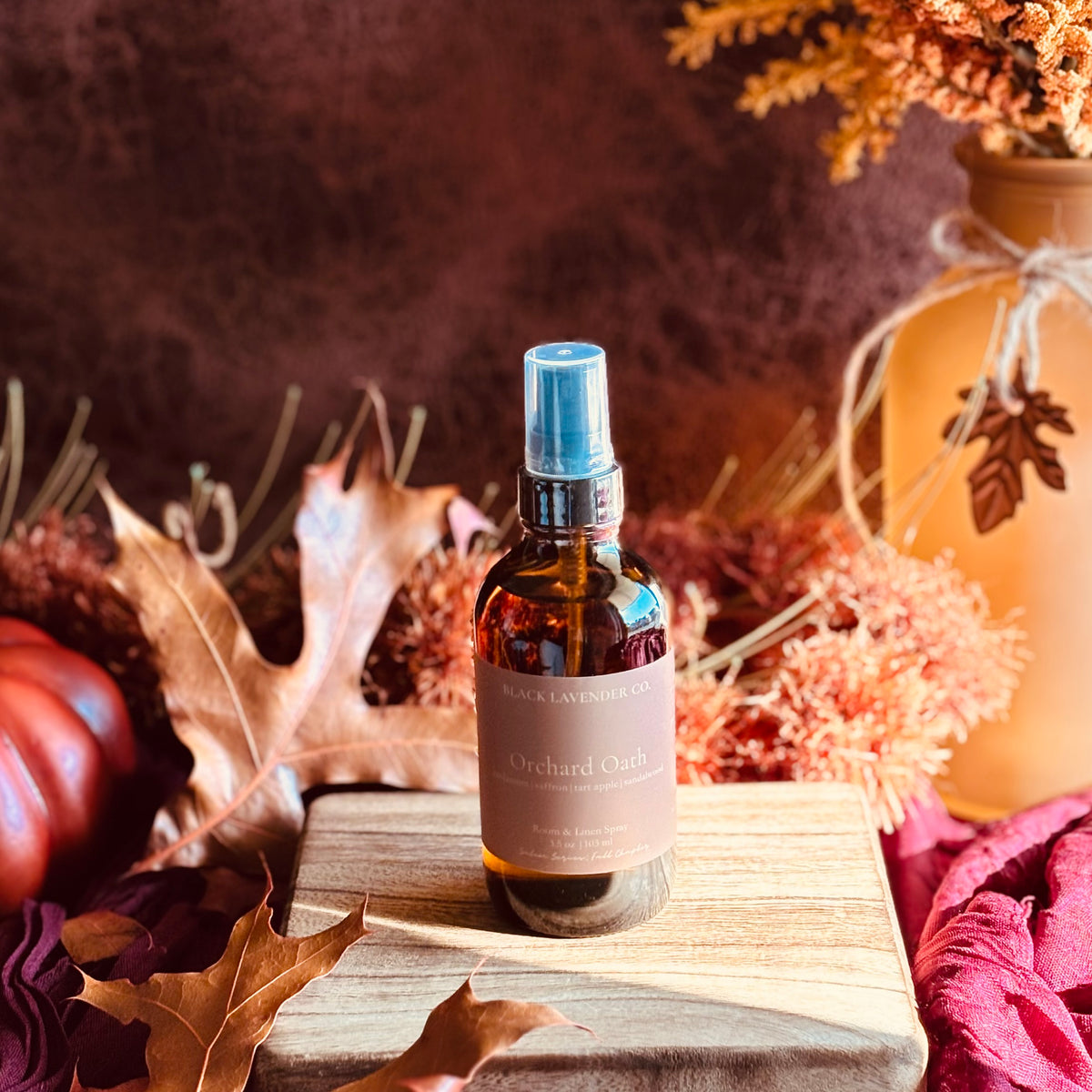 A bottle of Black Lavender Co. Orchard Oath room spray stands on a wooden block surrounded by autumn leaves, a small pumpkin, and dried floral decor.