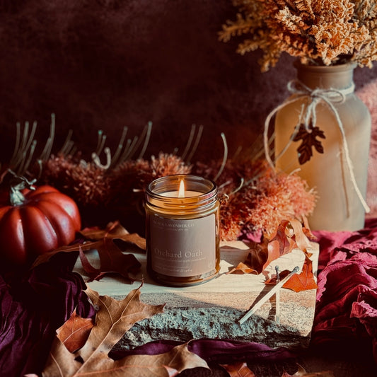 A lit candle in a amber glass jar sits on a wood slab surrounded by autumn leaves, dried flowers, a small red pumpkin, and a rustic beige vase against a dark warm background.