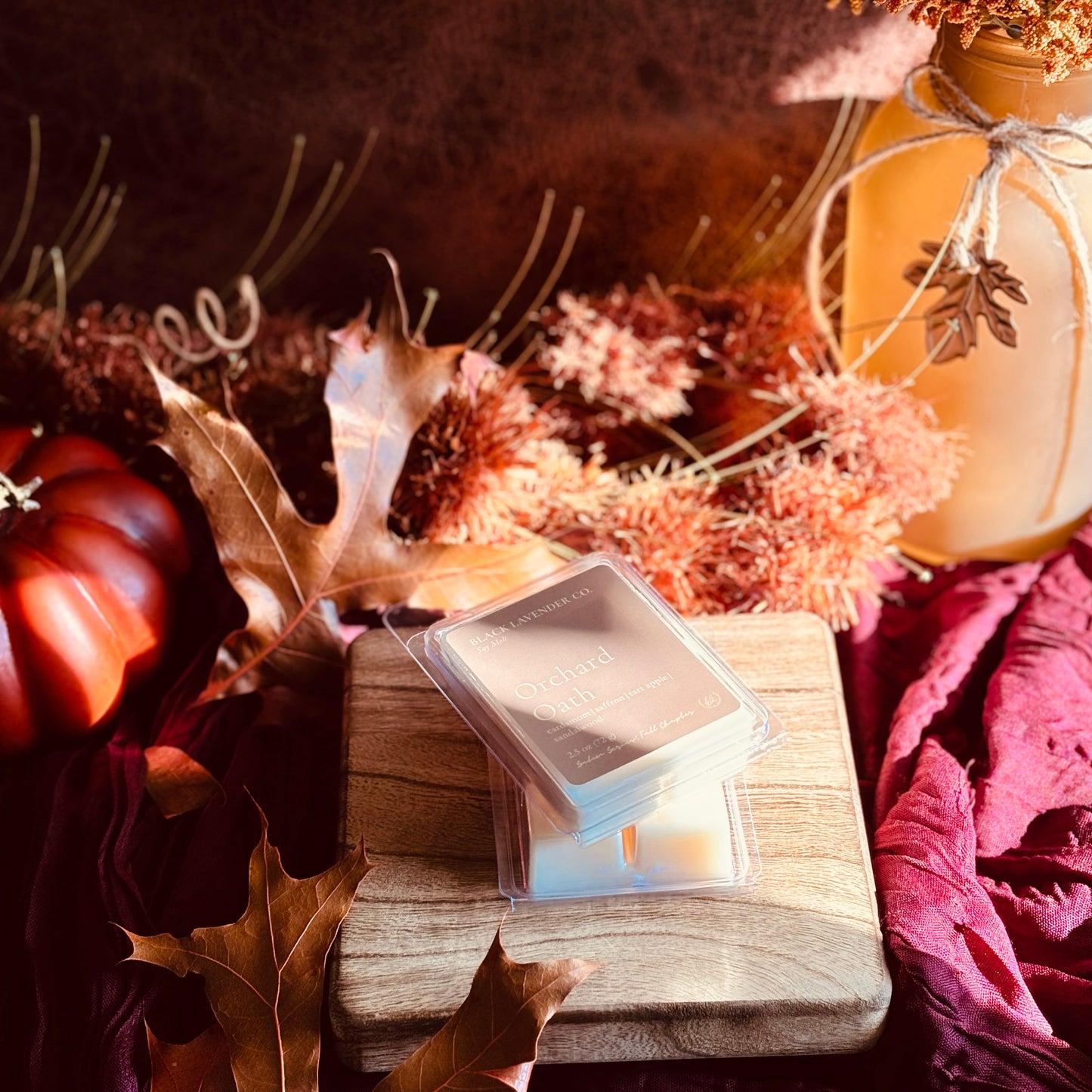 A stack of square wax melts in clear clamshell packaging sits on a wooden block, styled with autumn leaves, dried flowers, a small pumpkin, and a warm amber lantern in a cozy fall setting.