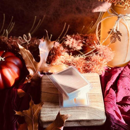A stack of square wax melts in clear clamshell packaging sits on a wooden block, styled with autumn leaves, dried flowers, a small pumpkin, and a warm amber lantern in a cozy fall setting.