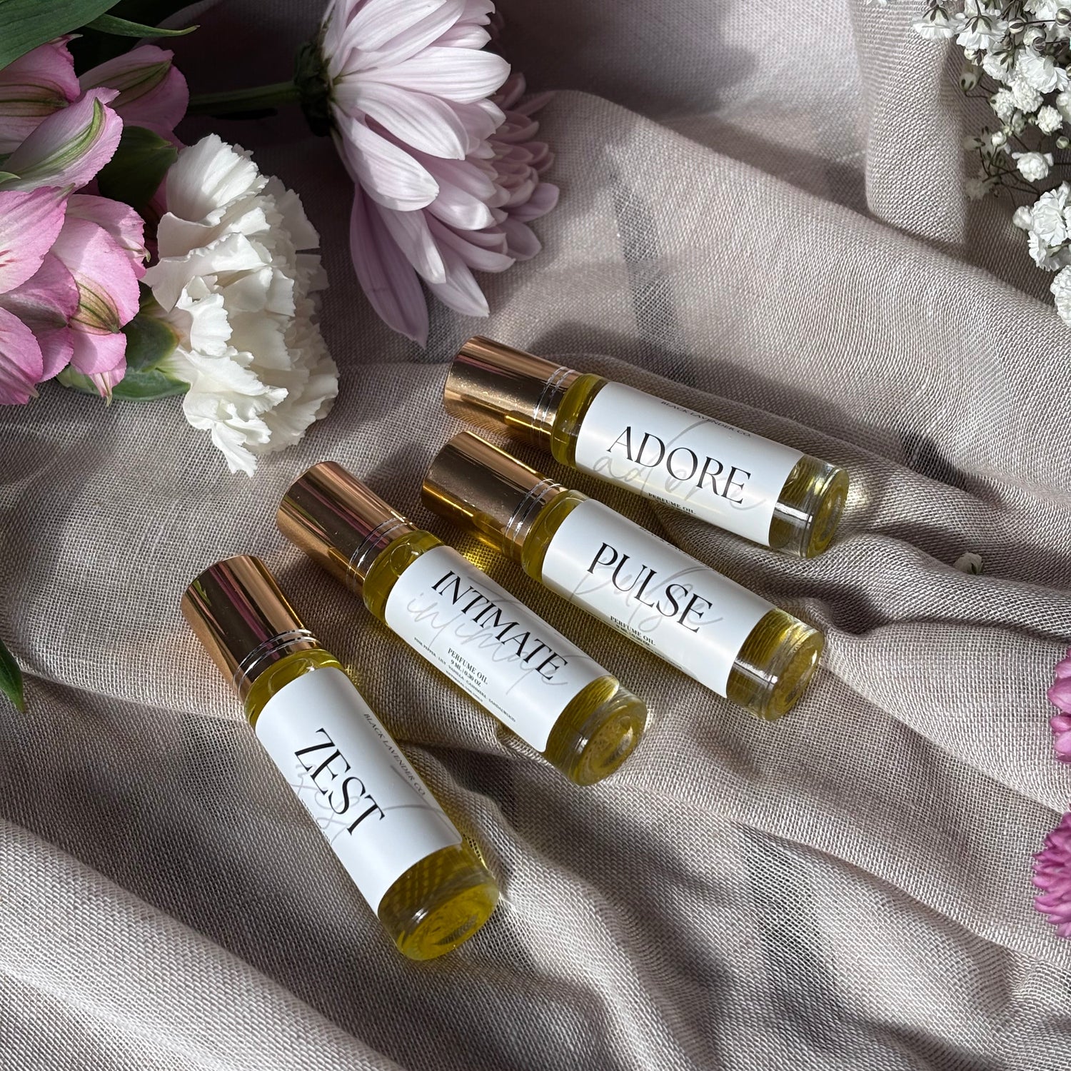 Roller bottles labeled 'Adore', 'Pulse', 'Intimate', and 'Zest' on a textured fabric background with flowers.