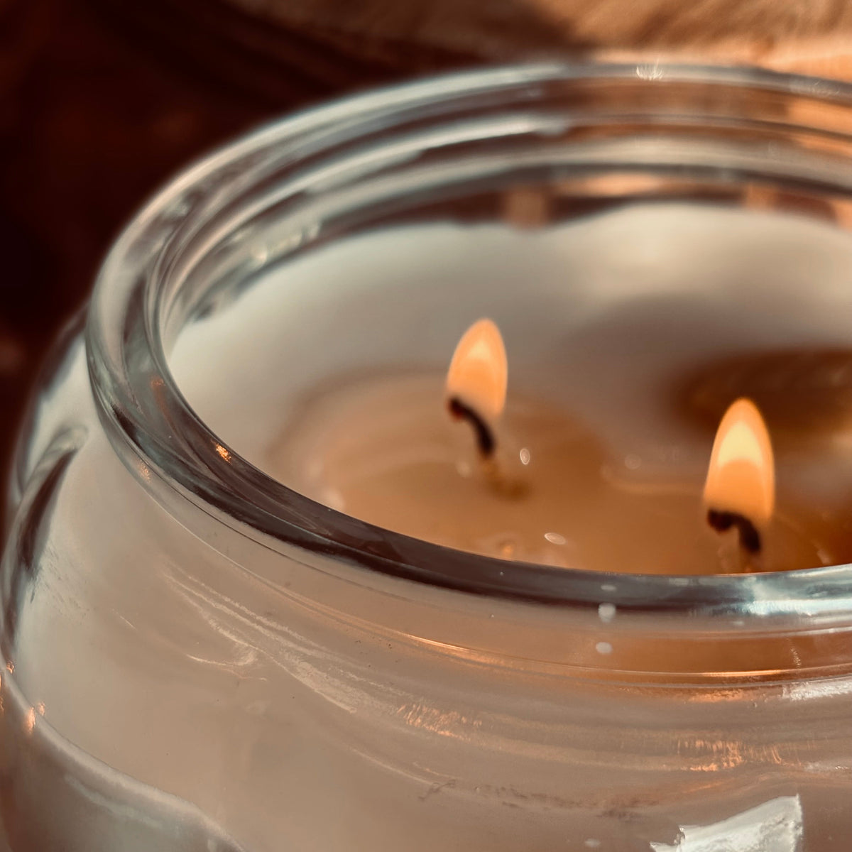 A close-up of a lit double-wick candle in a clear glass jar, showing two flames burning in warm amber tones against a dark brown background.