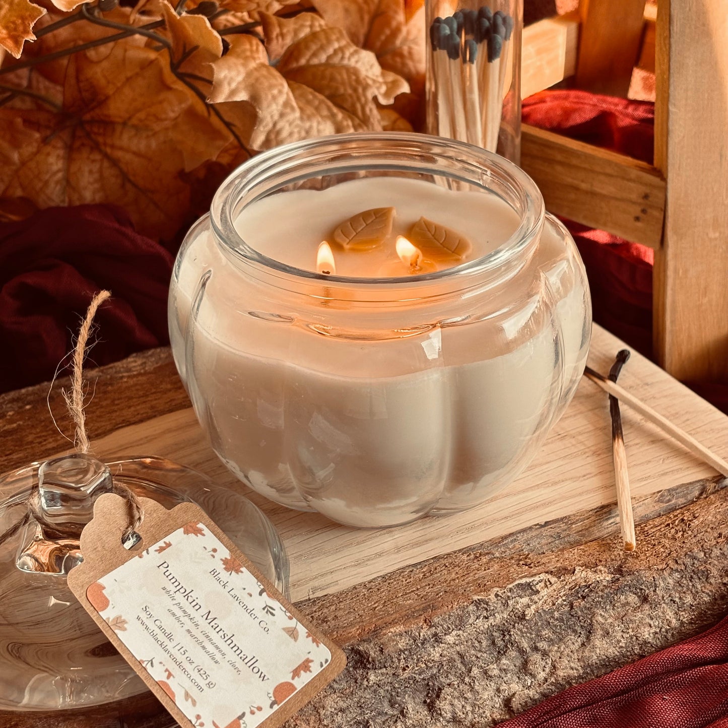 A lit glass jar candle with creamy off-white wax sits on a rustic wooden surface, surrounded by dried autumn leaves, red fabric, matches in a glass tube, and a small product tag reading 'Pumpkin Marshmallow'.