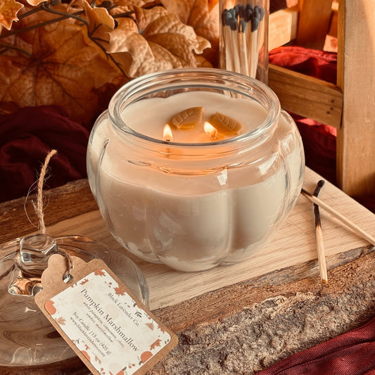 A lit glass jar candle with creamy off-white wax sits on a rustic wooden surface, surrounded by dried autumn leaves, red fabric, matches in a glass tube, and a small product tag reading 'Pumpkin Marshmallow'.