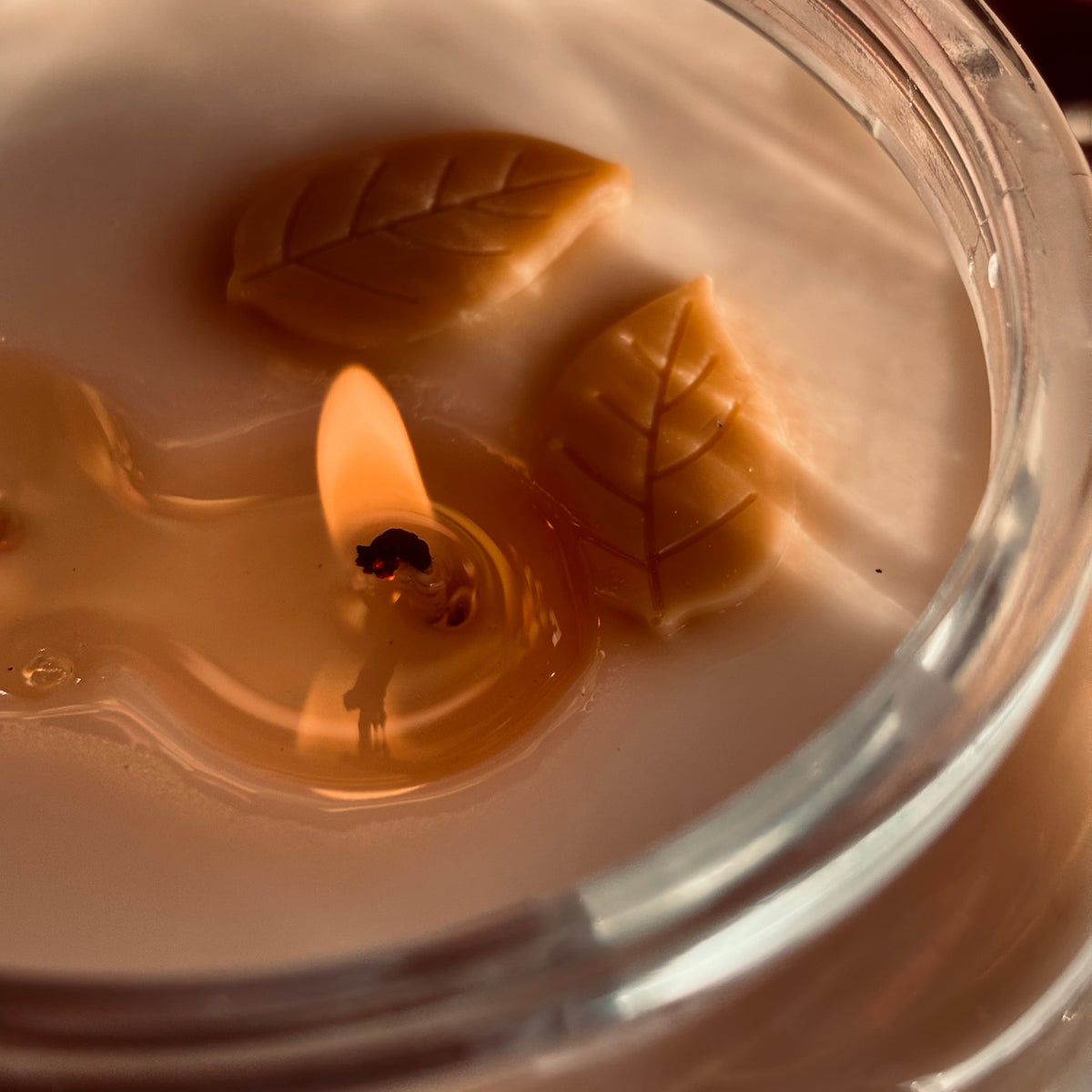 A close-up of a lit candle in a clear glass jar, showing creamy off-white wax, a small flame, and two decorative leaf shapes floating in the wax.