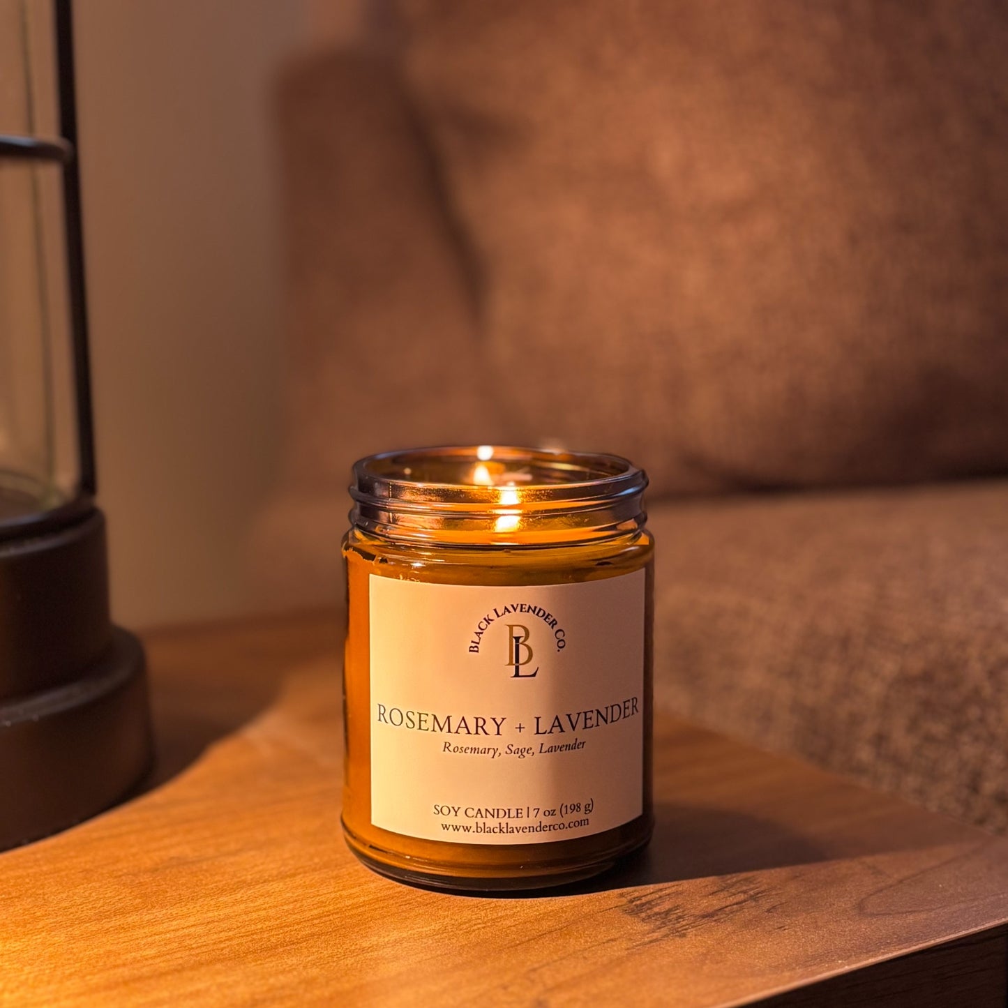 Candle in a glass jar labeled 'Rosemary + Lavender' on a wooden surface with a lifestyle background.