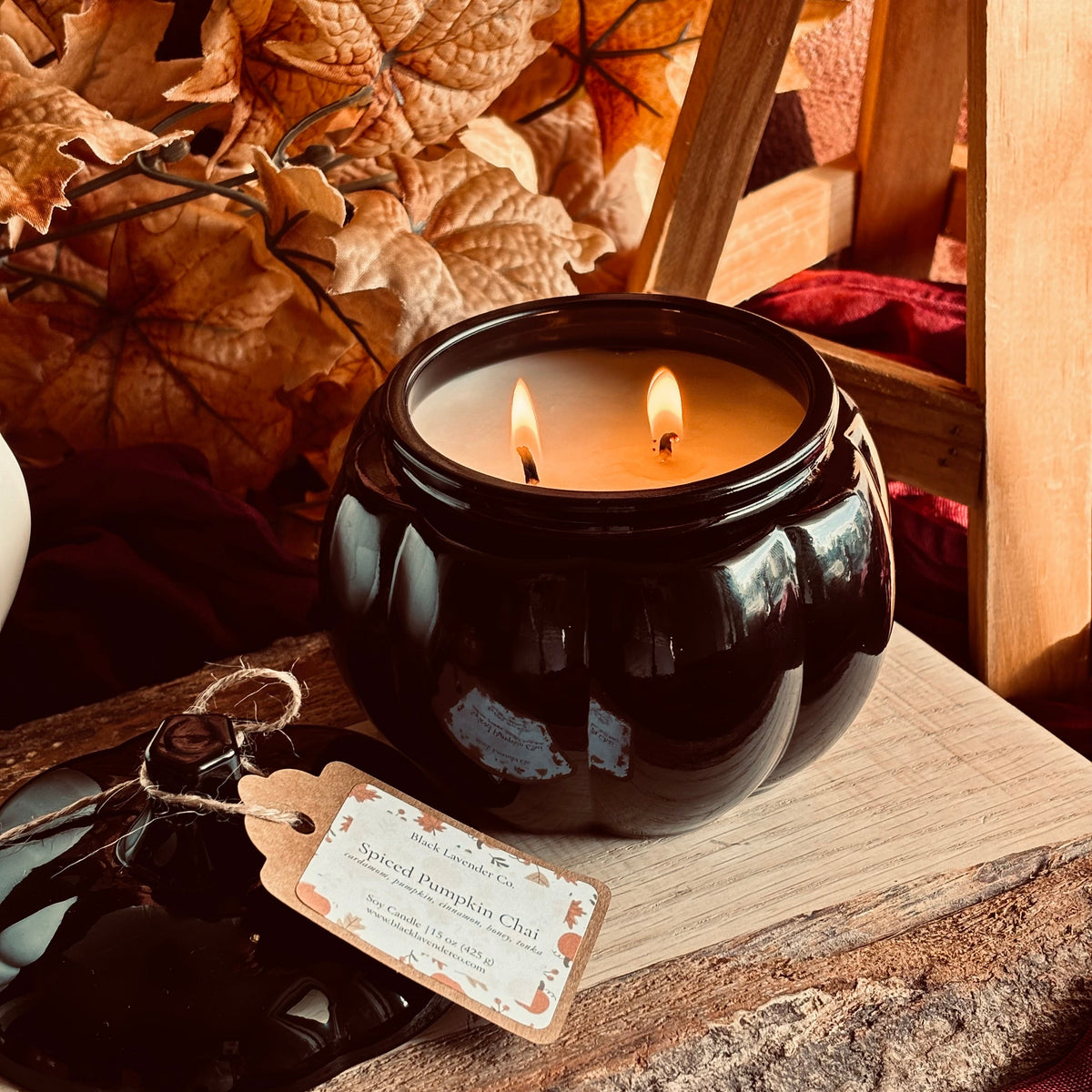 A lit black pumpkin-shaped candle jar with two wicks sits on a rustic wooden surface, surrounded by autumn leaves, wood, and burgundy fabric. 