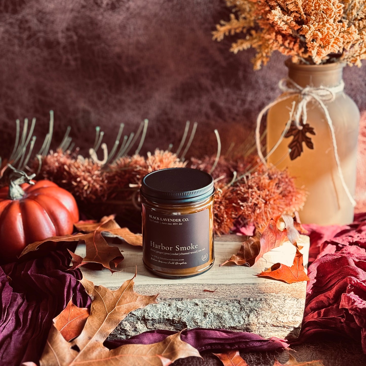 A amber jar candle labeled Harbor Smoke scented soy wax candle sits on a rustic wood slab surrounded by autumn leaves, a small orange pumpkin, dried flowers, and a decorative vase against a dark textured background.
