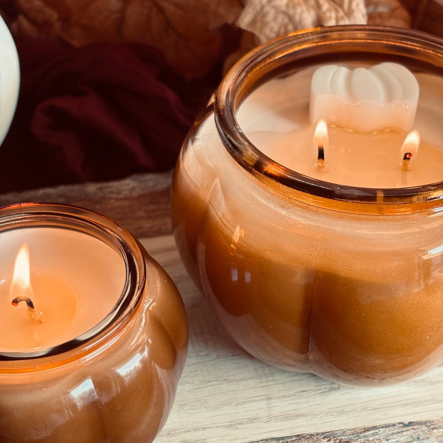 Two amber glass jar candles, one lit and one unlit, displayed on a rustic wooden surface with dried leaves in the background.