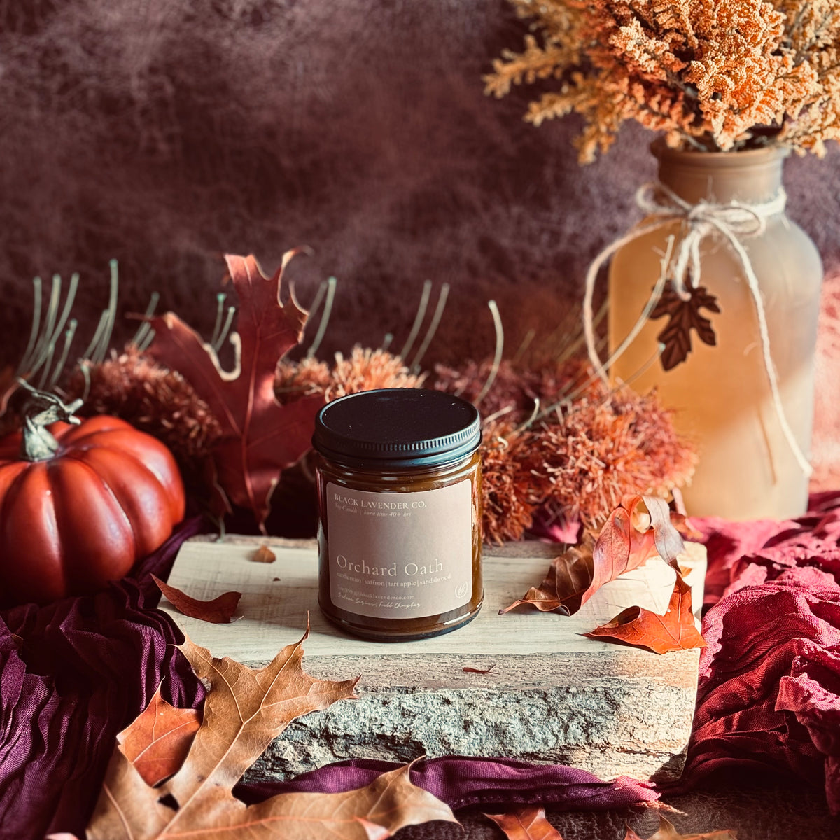 A jar candle labeled ' Orchard Oath' sits on a rustic wood slab surrounded by autumn leaves, dried flowers, and a small orange pumpkin in a styled fall scene.