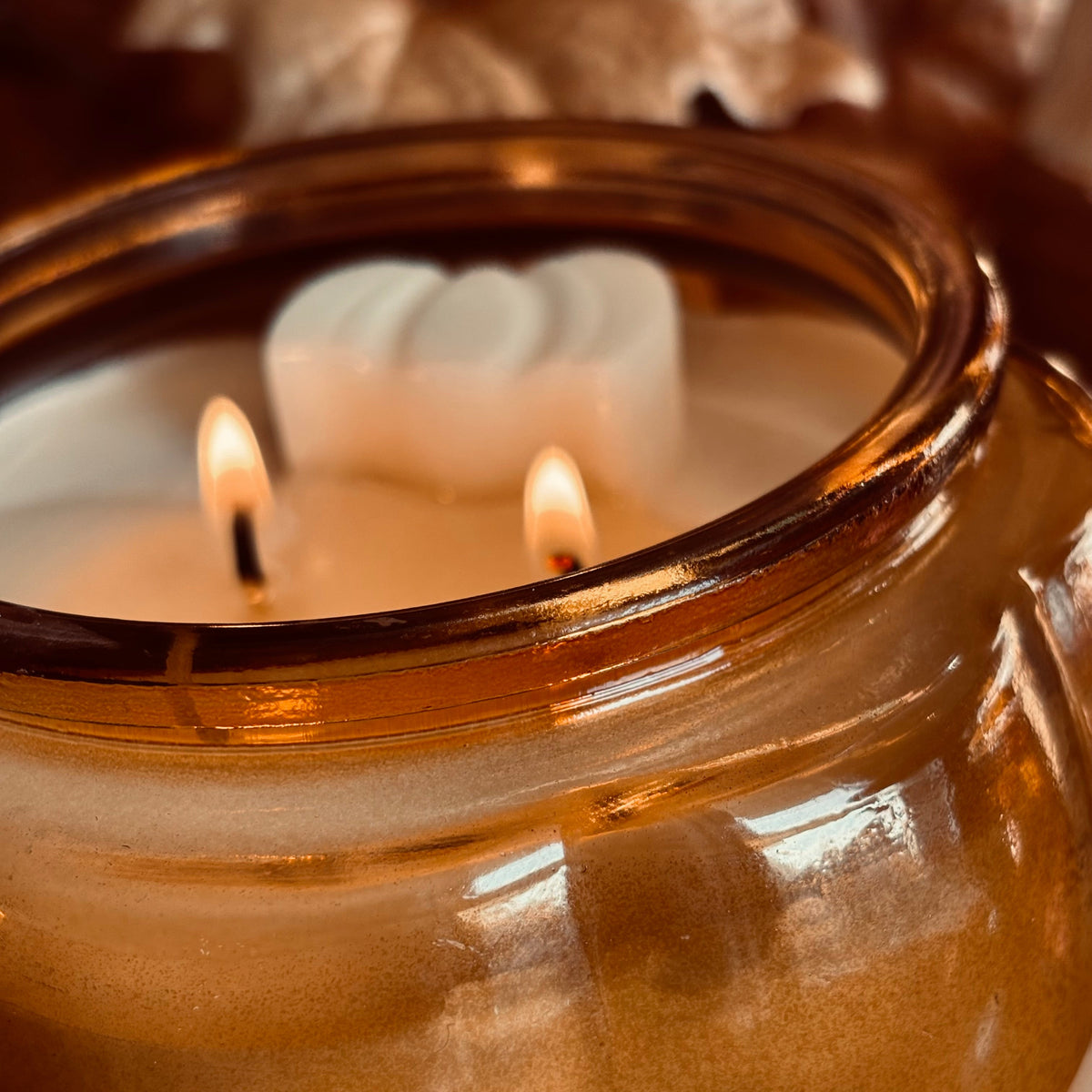 A close-up of a lit amber glass jar candle with two flames and a white wax insert, photographed in warm, cozy lighting.