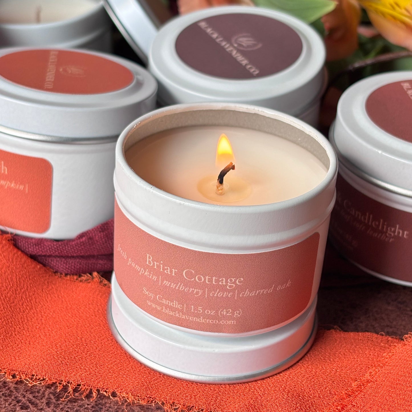 A lit tin candle labeled 'Briar Cottage' sits in the foreground with several matching candles around it on a textured orange fabric, with autumn leaves blurred in the background.
