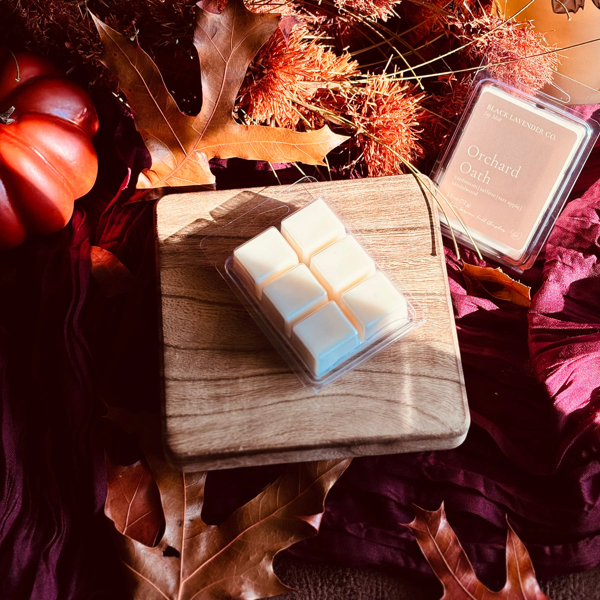 A wax melt product in a clear plastic clamshell container sits on a small wooden board, surrounded by autumn leaves, dried pumpkins, and fabric. The packaging label reads 'Black Lavender Co