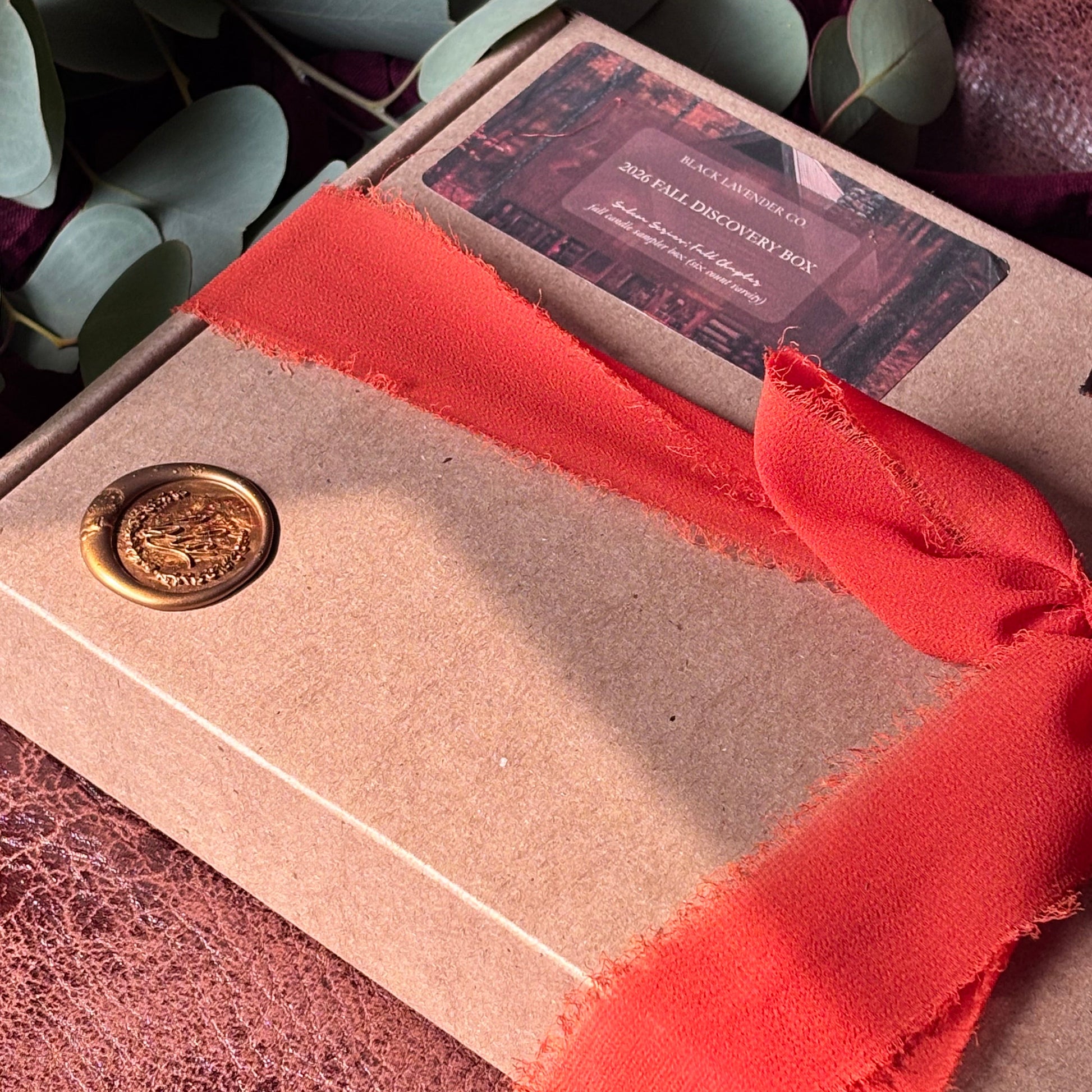A kraft paper gift box tied with a wide coral-red ribbon and sealed with a gold wax stamp, photographed from above on a textured mauve surface with eucalyptus leaves around it.