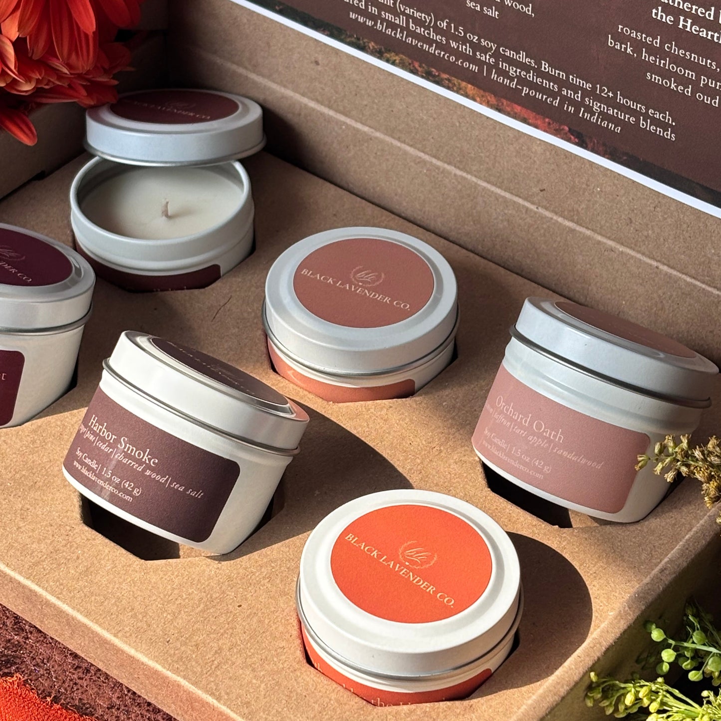 A curated collection of small white metal candles with white lids and labels in earthy tones, arranged inside a brown kraft gift box. One candle is shown open, revealing white wax.