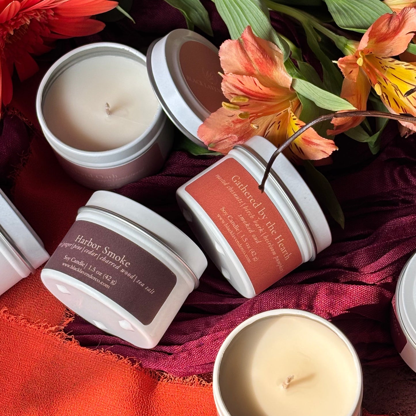 Several open white candle tins with labels are arranged on draped burgundy fabric with orange and peach flowers around them. Visible labels include 'Harbor Smoke' and 'Gathered by the hearth'