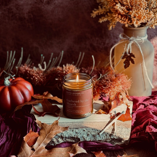 A lit candle in a amber glass jar sits on a rustic wooden slab surrounded by autumn leaves, dried flowers, a small red pumpkin, and a beige vase with twine in a moody fall-themed setting.
