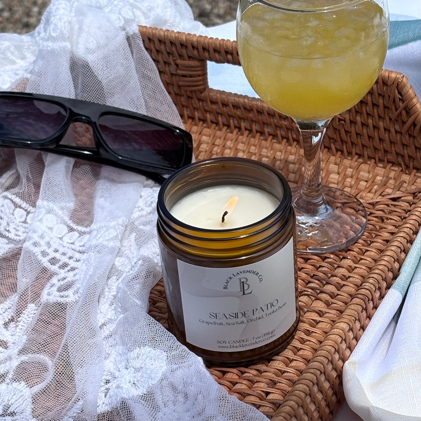 Candle labeled 'Seaside Patio' on a woven tray with sunglasses and a glass of yellow liquid.